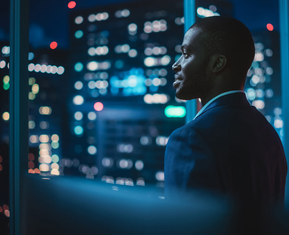A businessman working late and staring out into a lit-up cityscape.