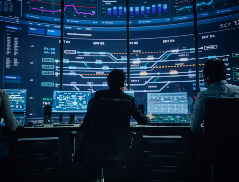 Team of analysts monitoring multiple digital dashboards and data visualizations on large screens in a control room.