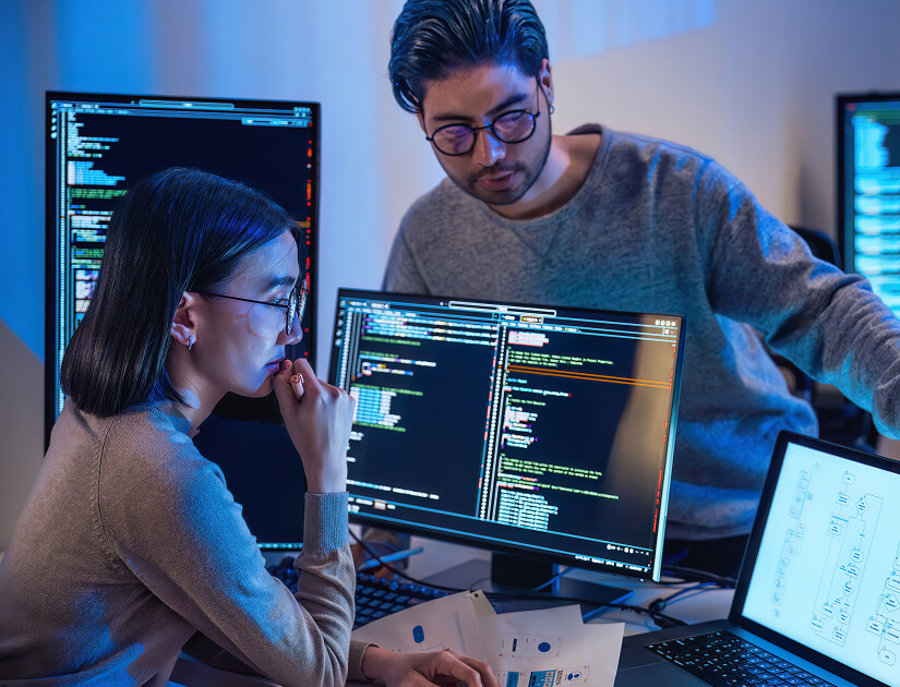 Two software developers reviewing code on multiple monitors in an office setting.
