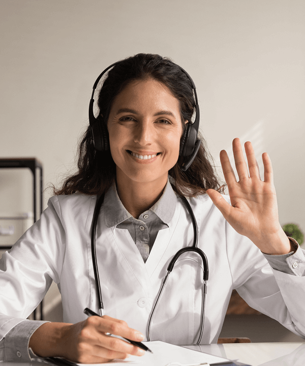 Smiling doctor wearing headphones and a stethoscope, waving during an online telehealth appointment.