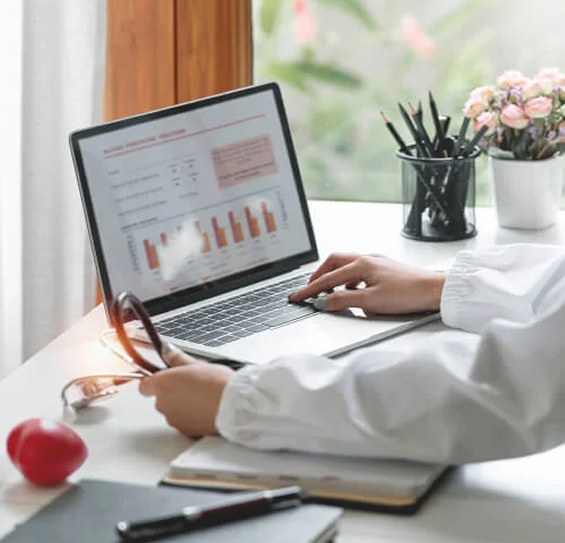 Person in a white coat working on a laptop displaying charts and graphs at a desk with notebooks, pens, and flowers nearby.