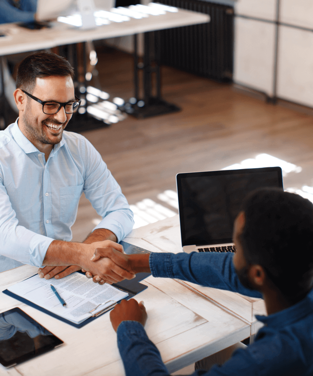 Two business professionals smiling and shaking hands across a desk with a laptop and documents.